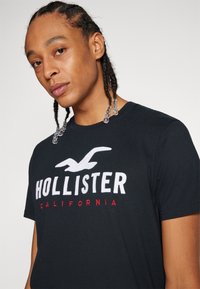 Melnas kokvilnas T-krekls ar "Hollister" logotipu baltā un sarkanā izšuvumā, apaļa apkakle, īsas piedurknes un gluda tekstūra.