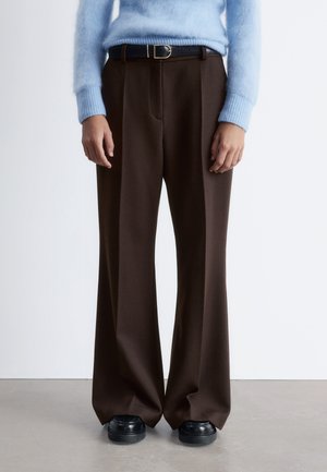 Braune Bundfaltenhose aus einem weichen, strukturierten Stoff, kombiniert mit einem hellblauen Pullover und schwarzem Schuhwerk.