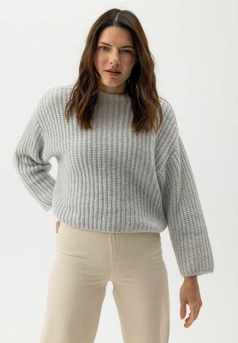 Szürke, bordázott kötött pulóver kerek nyakkal és hosszú ujjakkal, világos bézs nadrággal párosítva. Az anyag puhának és texturáltnak tűnik.