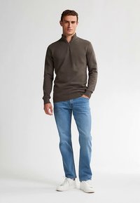 Maglione marrone scuro con zip a quattro quarti e motivo a rilievo, abbinato a jeans azzurri e sneakers bianche. Modello in piedi davanti a uno sfondo chiaro.