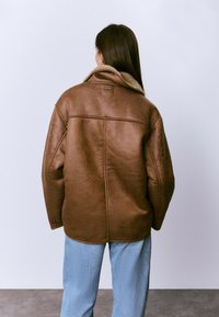 Mujer con cabello largo, vistiendo una chaqueta de piel de oveja marrón y jeans de color azul claro, de pie frente a una pared blanca lisa.