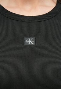 Svart ribbet tanktopp med en bred halslinning. Den har en rektangulær Calvin Klein-logo nær midten. Myk stofftekstur.