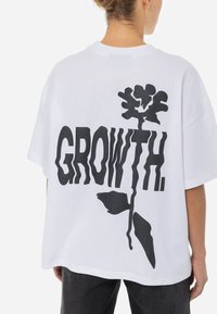 Persona vistiendo una camiseta blanca de gran tamaño con un gráfico negro de una flor y la palabra "CRECIMIENTO" en la parte trasera, vista desde atrás.