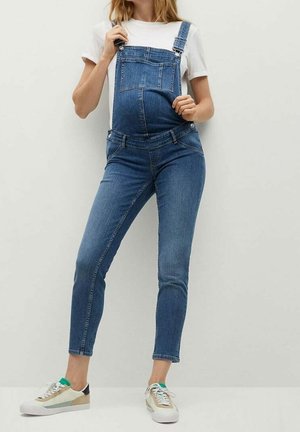 Femme enceinte portant une salopette de maternité en denim bleu, un t-shirt blanc et des baskets, debout devant un fond uni.