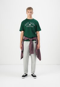 Grønn bomull T-skjorte med "GAP 1968 ATHLETICS" trykk, grå joggebukser, og ruterød og svart skjorte knyttet rundt livet. Svarte sneakers.