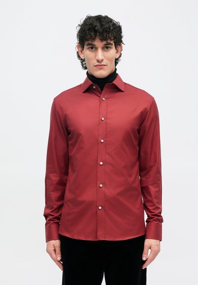 Burgundijska košulja dugih rukava s glatkom teksturom, dugmadima sprijeda i uskim ovratnikom, nosi se preko crnog rolka.