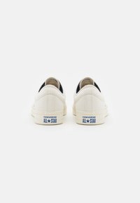 Bijele platnene tenisice s plavim "CONVERSE ALL STAR" brandingom na peti, sa zaobljenim oblikom i teksturiranim gornjim dijelom od tkanine.
