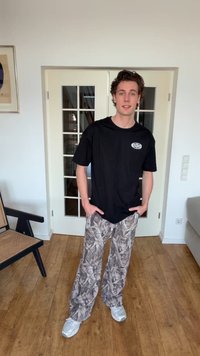 Νεαρός άνδρας που στέκεται μέσα σε κλειστό χώρο φορώντας μαύρο μπλουζάκι Racing Club, φαρδιά παντελόνια με σχέδια και λευκά αθλητικά παπούτσια, με τα χέρια στις τσέπες.