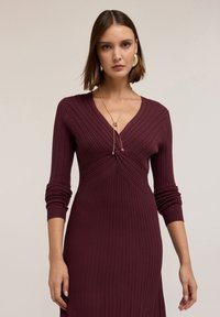 Abito a maglia ribbed bordeaux con maniche lunghe, scollatura profonda a V e dettaglio frontale annodato, caratterizzato da una sottile texture e una silhouette aderente.