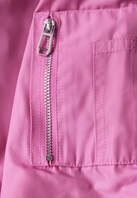 Różowy materiał z diagonalną teksturą, srebrny zamek błyskawiczny po prawej stronie oraz kieszeń naszywaną z detalami w szyciu po lewej stronie.