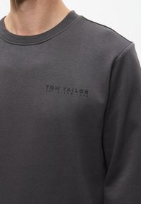 Ciemnoszary sweatshirt z okrągłym dekoltem i subtelnym wytłoczonym logo na piersi. Miękka tkanina o gładkiej fakturze oraz długie rękawy.