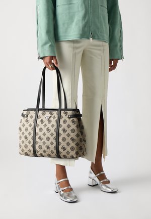 Femme tenant un sac fourre-tout beige et noir à motifs, portant une veste vert menthe, un pantalon crème à fente à l'ourlet et des escarpins argentés Mary Jane.