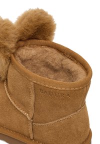 Brązowy zamszowy botek za kostkę z miękką beżową futrzaną wyściółką, ozdobiony okrągłym pomponem na górze oraz wzmocnionym przeszyciem wzdłuż szwów.