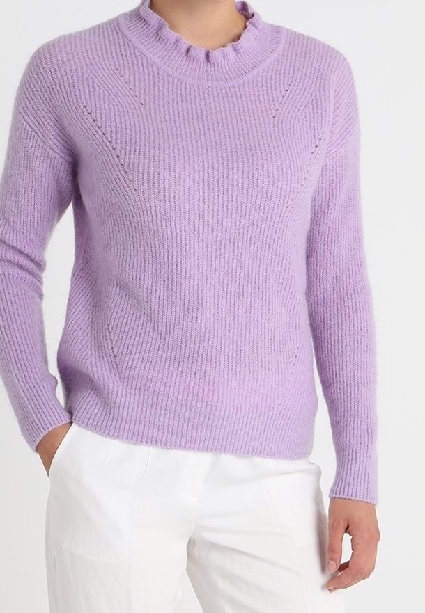 Personne portant un pull en maille lavande à manches longues avec un col volanté et un pantalon blanc, debout avec une main dans la poche.