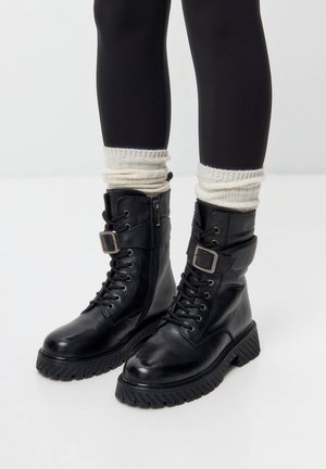 Black leather ankle boots with a thick rubber outsole, lace-up front, side zipper, and adjustable strap with a metal buckle.