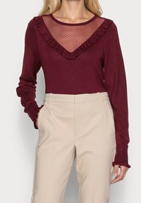 Burgundy långärmad topp med en genomskinlig prickig mesh-krage och volangkanter, tillsammans med krämfärgade högmidjade byxor.