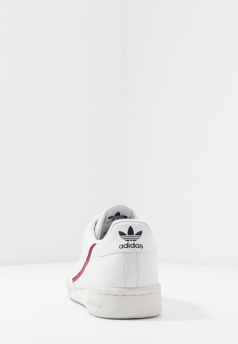 White athletic shoe with a red and blue stripe, rubber sole, and a textured panel. Adidas logo displayed on the heel and tongue.