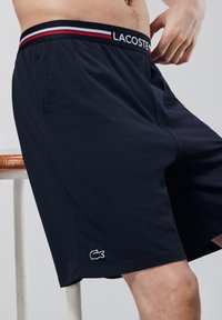 Bermuda blu in tessuto morbido, con una cintura a righe bianche, nere e rosse e un piccolo logo bianco Lacoste sul bordo.