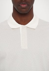Gros plan d'un homme portant un polo blanc avec un motif géométrique gris discret et un col blanc uni.