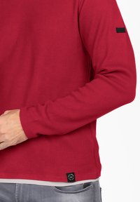 Chemise longue à manches en tricot gaufré rouge avec un col rond et des poignets côtelés. Présente une étiquette noire sur le côté inférieur gauche.