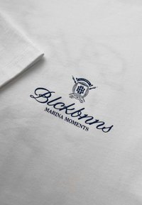 Witte katoenen t-shirt met een marineblauw geborduurd logo en de tekst "Blockanns MARINA MOMENTS" met nautisch embleem detail.