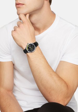Montre noire avec un boîtier rond en argent, un cadran noir avec des accents et des marquages jaunes, sur un bracelet en cuir noir avec des coutures jaunes.