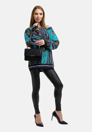 Kvinde iført en farverig mønstret sweater, sorte læderleggings, sorte højhælede sko, bærende på en sort håndtaske med kæderem.