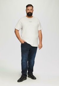 Jasnoszara koszulka z krótkim rękawem z logo, zestawiona z ciemnoniebieskimi dżinsami i czarnymi sneakersami. Model stoi z jedną ręką w kieszeni.
