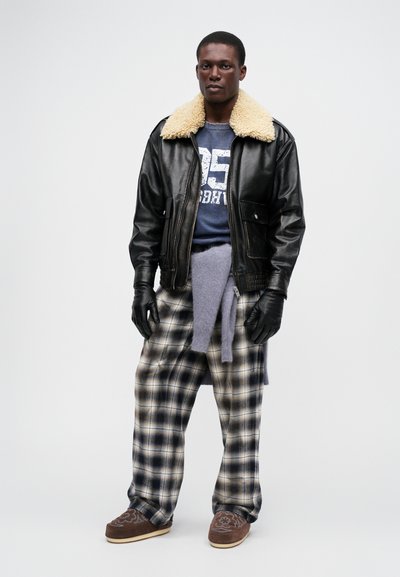 Veste en cuir noir avec col en shearling, sweat gris avec imprimé, pantalon à carreaux, chaussures marron à motifs et gants noirs.