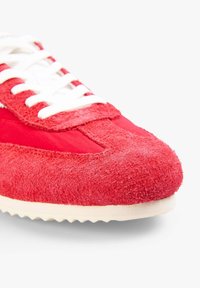 Zapatilla roja que combina ante y tejido. Cierre de cordones blancos y puntera texturizada y ligeramente elevada. Suela exterior de goma suave con tracción.