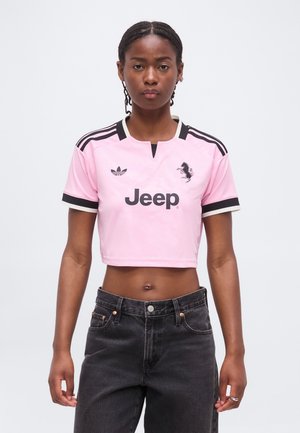 Mujer negra con el pelo trenzado que lleva una camiseta deportiva rosa corta con rayas negras, logo de Jeep y jeans negros de talle alto.