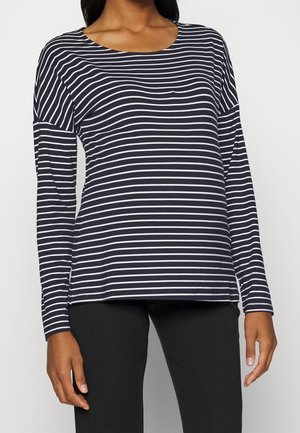 Zwangere vrouw die een zwart-wit gestreept shirt met lange mouwen en zwarte broek draagt, staand tegen een effen achtergrond.