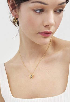 Ženska, ki nosi zlata uhančka v obliki čebel in ujemajoče se ogrlico z obeskom v obliki čebele, oblečena v belo majico, gleda navzdol in v desno.