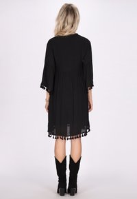 Schwarzes Kleid mit langen Glockenärmeln, geraffter Taille und fransigem Saum. Knielange Länge, aus leichtem Stoff gefertigt. Hochhackige schwarze Stiefel.