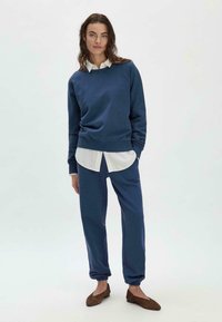 Felpa blu navy e pantaloni coordinati in tessuto morbido, con polsini e cintura a coste. Camicia bianca visibile sotto la felpa.