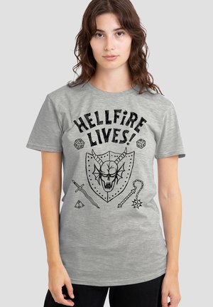 Junge Frau trägt ein graues T-Shirt mit dem Schriftzug "HELLFIRE LIVES!" und schwarzen Fantasy-Motiven, darunter ein gehörnter Totenschädel, Würfel, Schwert und Streitkolben.