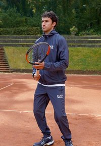 Navy athletic tracksuit with zipped hood, logo on chest, and elastic cuffs. Holding a tennis racket on a clay court. Brown background.