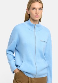 Jasnoniebieski sweter zapinany na zamek wykonany z miękkiego materiału. Posiada wysoki kołnierz oraz małą kieszeń na piersi z srebrnym zamkiem. Luźny krój z bocznymi kieszeniami.