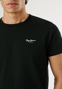 Junger Mann trägt ein schwarzes Pepe Jeans London T-Shirt mit kurzen Ärmeln und Rundhalsausschnitt, dargestellt von den Schultern bis zur Mitte des Oberkörpers.