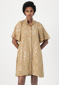 Abito color tan con maniche corte e volants e colletto alto. Presenta un motivo astratto in argento lucido su tutta la superficie. Il tessuto leggero si drappeggia in modo elegante.
