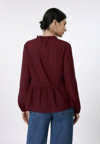 Μπορντό μπλούζα με γιακά με βολάν, μακριά μανίκια και τελείωμα πεπλούμ· απαλό ύφασμα· συνδυασμένη με μπλε τζιν παντελόνι· λεία υφή.