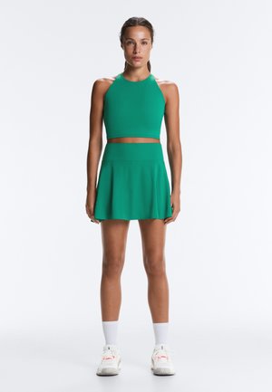 Kvinde står med front mod kamera iført en grøn ærmeløs crop top og matchende nederdel, hvide sokker og hvide sneakers på en hvid baggrund.