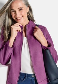 Veste ajustée en matériau velouté doux couleur violet clair avec un col haut, fermeture éclair, associée à un sac à main bleu marine et un haut clair.