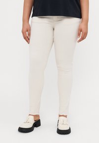 Világos bézs skinny farmernadrág, rojtos szegéllyel, denim szövetből készült. Fehér cipővel párosítva, amelyet frufru részlet és vastag fekete talp jellemez.