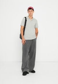 T-shirt polo gris, pantalon large gris, sandales noires et une casquette rouge. Les pantalons sont amples, avec une texture douce et des détails subtils.
