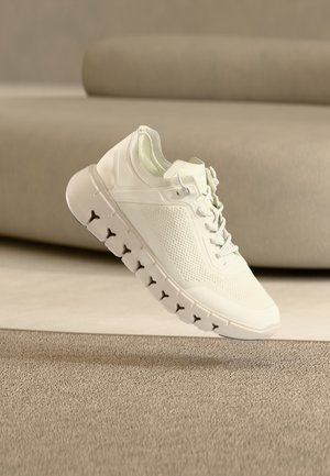 Chaussure de sport blanche avec une tige en mesh texturé, une semelle coussinée, un design à lacets et une semelle rainurée segmentée, présentée sur un fond neutre.