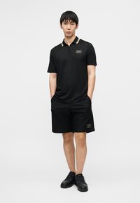 Polo noir à manches courtes avec un logo accentué d'or, assorti à un short en mesh noir et des chaussures noires, présentant un design décontracté.