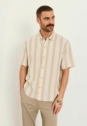 Mann mit Schnurrbart, der ein beige-weiß gestreiftes Hemd mit kurzen Ärmeln und beige Hosen trägt, steht mit den Händen in den Taschen.