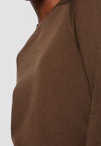 Gros plan d'une personne portant un pull marron à manches longues en tricot, avec des détails de coutures visibles à l'épaule et à la manche.