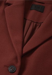 Ruosteenvärinen villablazer pystyselällä, jossa on lovettu kaulus, yksittäinen musta nappikiinnitys ja sileä satiinivuori. Sisältää hienovaraisen tekstuurin.